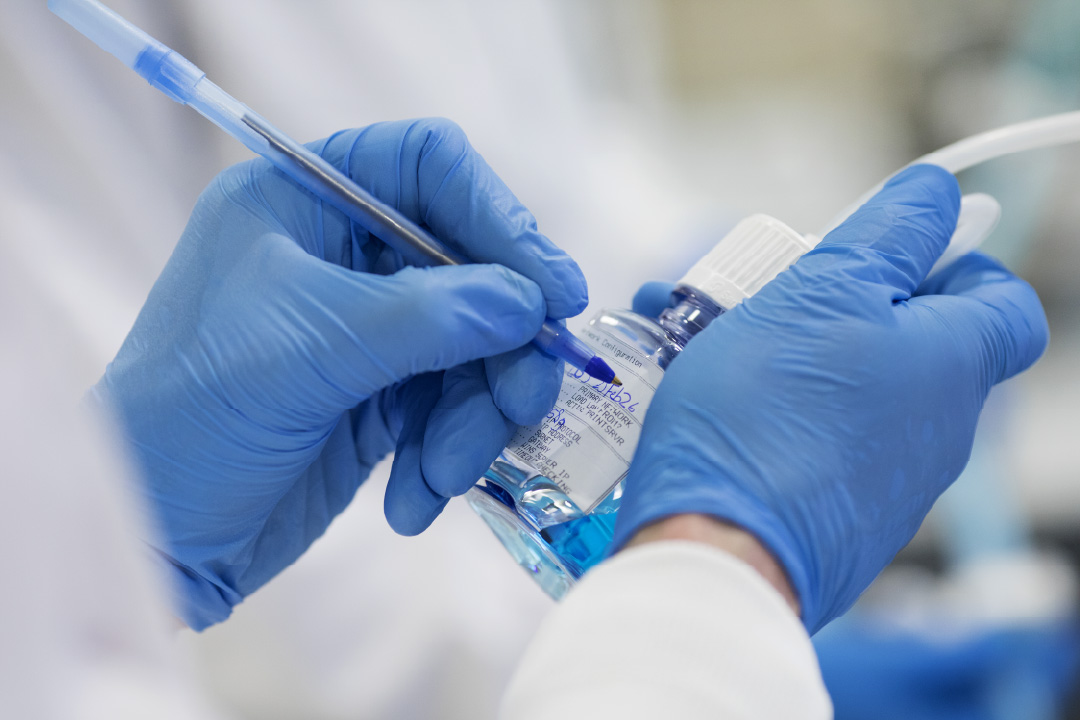 Gowned technician in blue gloves handling SaniSure PharmaTainer bottles connected to a multi-port sampling manifold in a controlled manufacturing environment
